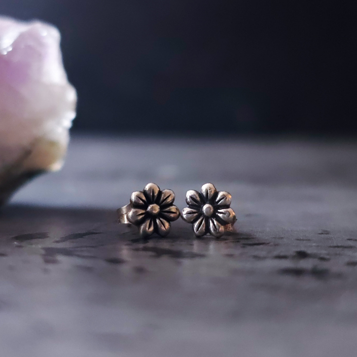 Pair of silver flower-shaped earrings on a dark surface with a blurred purple crystal in the background.