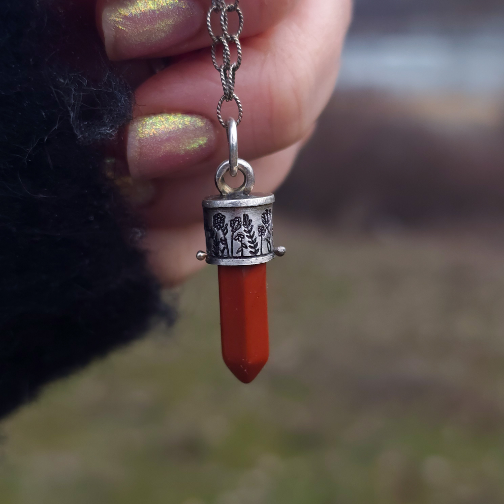 Hand holding a pendant with a red jasper and silver chain against a blurred natural background
