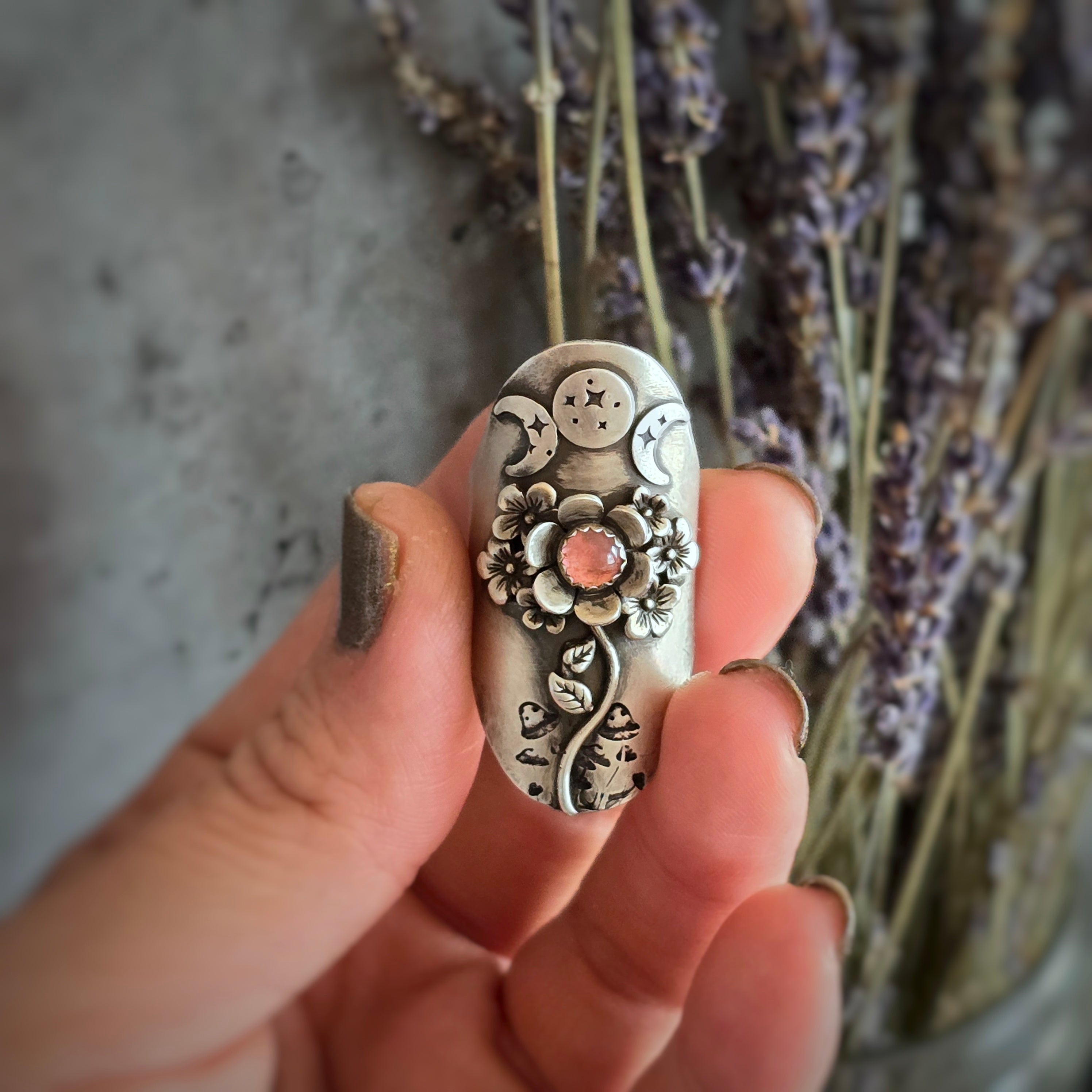 The image shows a hand holding a unique, intricately designed silver ring depicting a goddess figure adorned with floral and celestial elements. The ring features a pink tourmaline stone as the centerpiece, resembling a blooming flower. Surrounding the central stone are delicate cherry blossom flowers and triple moon phase symbols, adding to the ring's ethereal and mystical aesthetic.