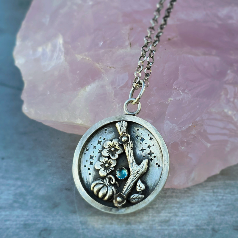 Sterling Silver Pumpkin Branch cherry blossoms round pendant with a small London Blue topaz stone displayed in front of a rose quartz crystal 